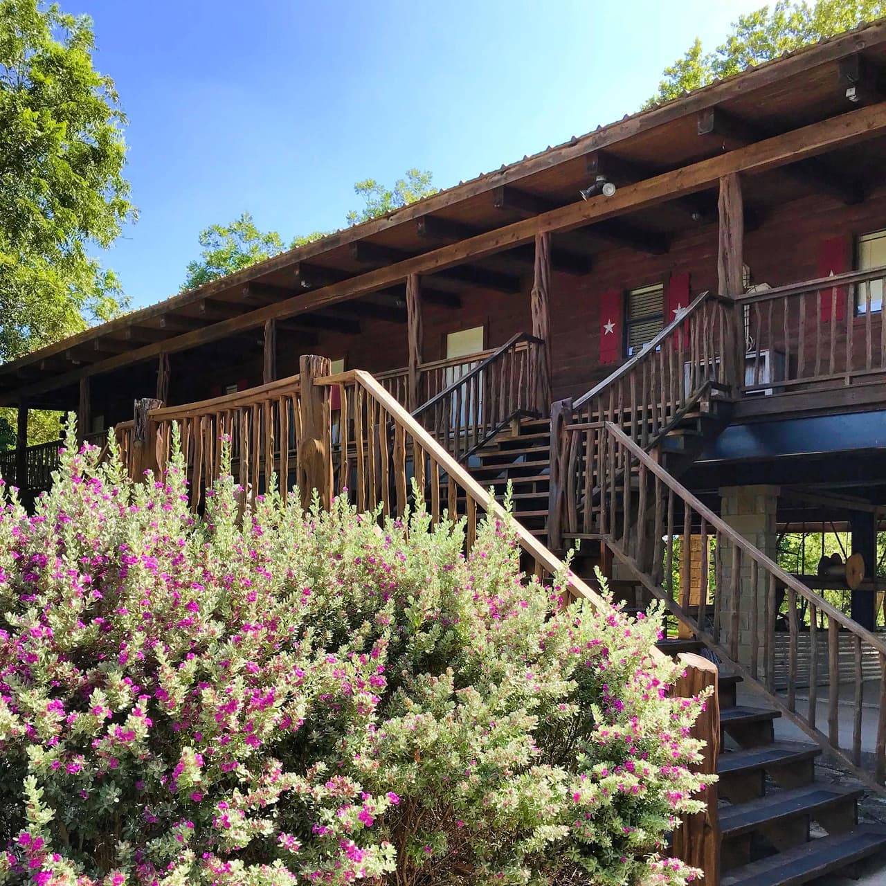 Sabinal River Lodge exterior with red window shutters, log-post stairs, and blooming crepe myrtle shrubs