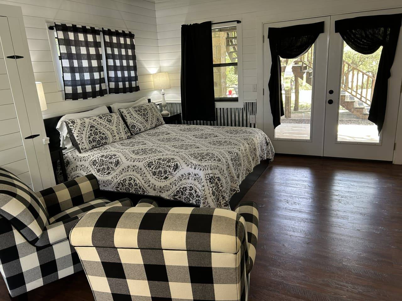 Sabinal Suite bedroom at Sabinal River Lodge in Utopia, Texas, with a black bed frame, floral bedspread, buffalo-check armchairs, and French doors to the patio