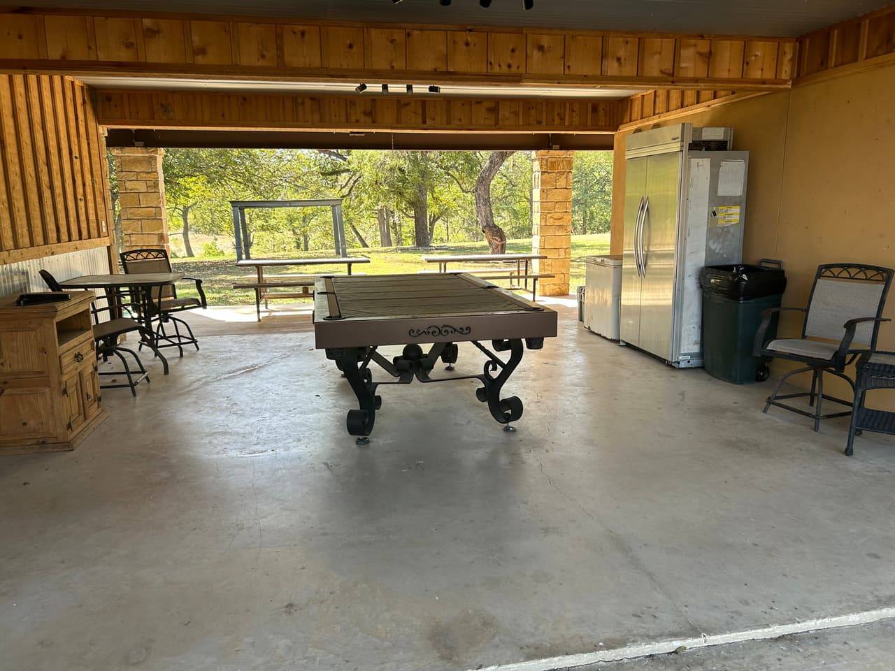 Covered outdoor recreation pavilion at Sabinal River Lodge in Utopia, Texas, with a pool table, patio seating, and picnic tables overlooking the lawn